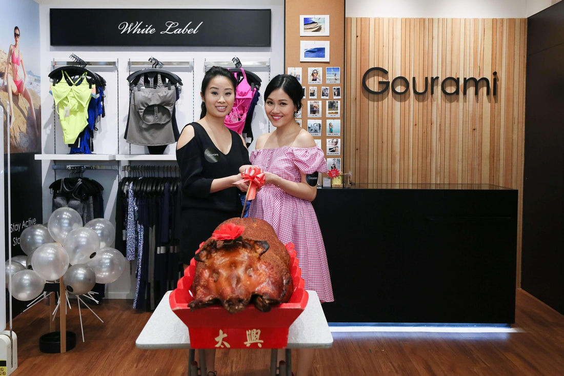 Two women at a swimwear and activewear store ribbon-cutting ceremony with a roast pig centerpiece