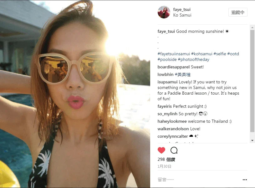 Woman in sunglasses and palm print swimwear taking a poolside selfie in sunny outdoor setting.