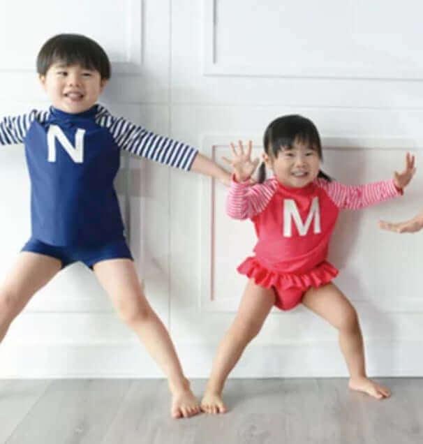 Two kids in colorful swimsuits posing playfully indoors, boy in blue, girl in red ruffle suit