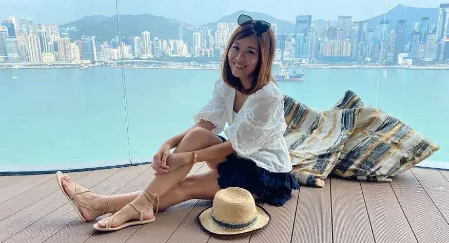 Woman in casual summer outfit sits by waterfront with city skyline and hat on wooden deck