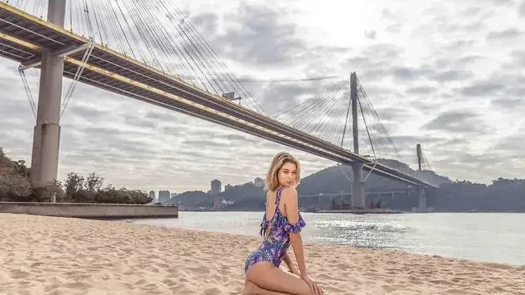 Woman in a purple floral one-piece swimsuit posing on a sandy beach near a large suspension bridge