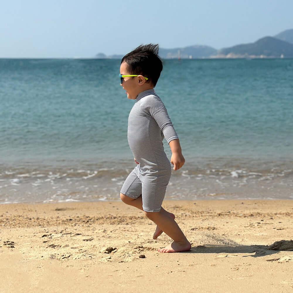 Young boy in a gray swimsuit running on the beach with sunglasses, enjoying a sunny day by the water.