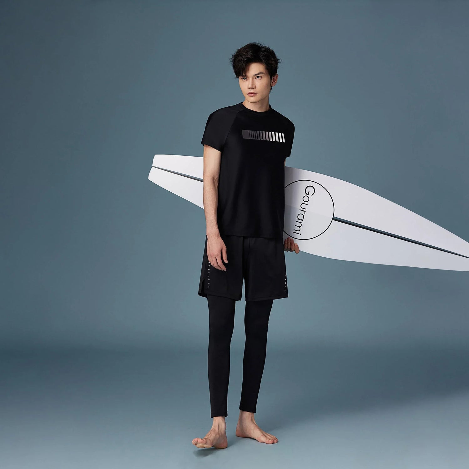 Stylish male model in black surfwear holding a surfboard, showcasing a modern athletic collection for men.