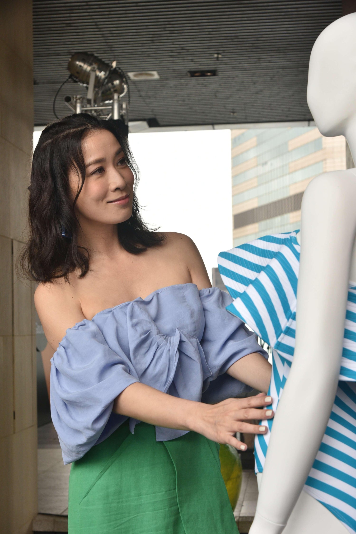 Woman in off-shoulder blue blouse interacting with a striped mannequin outdoors.