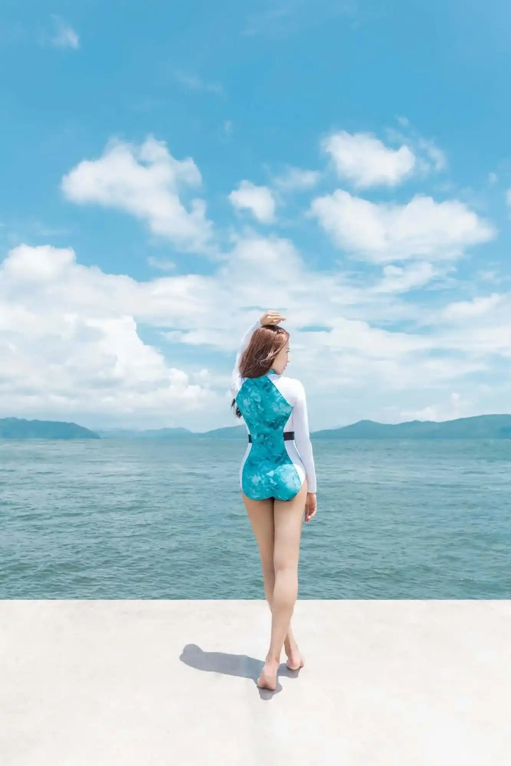 一名女子穿着蓝白相间的长袖连体泳衣，站在蓝天下的海边。