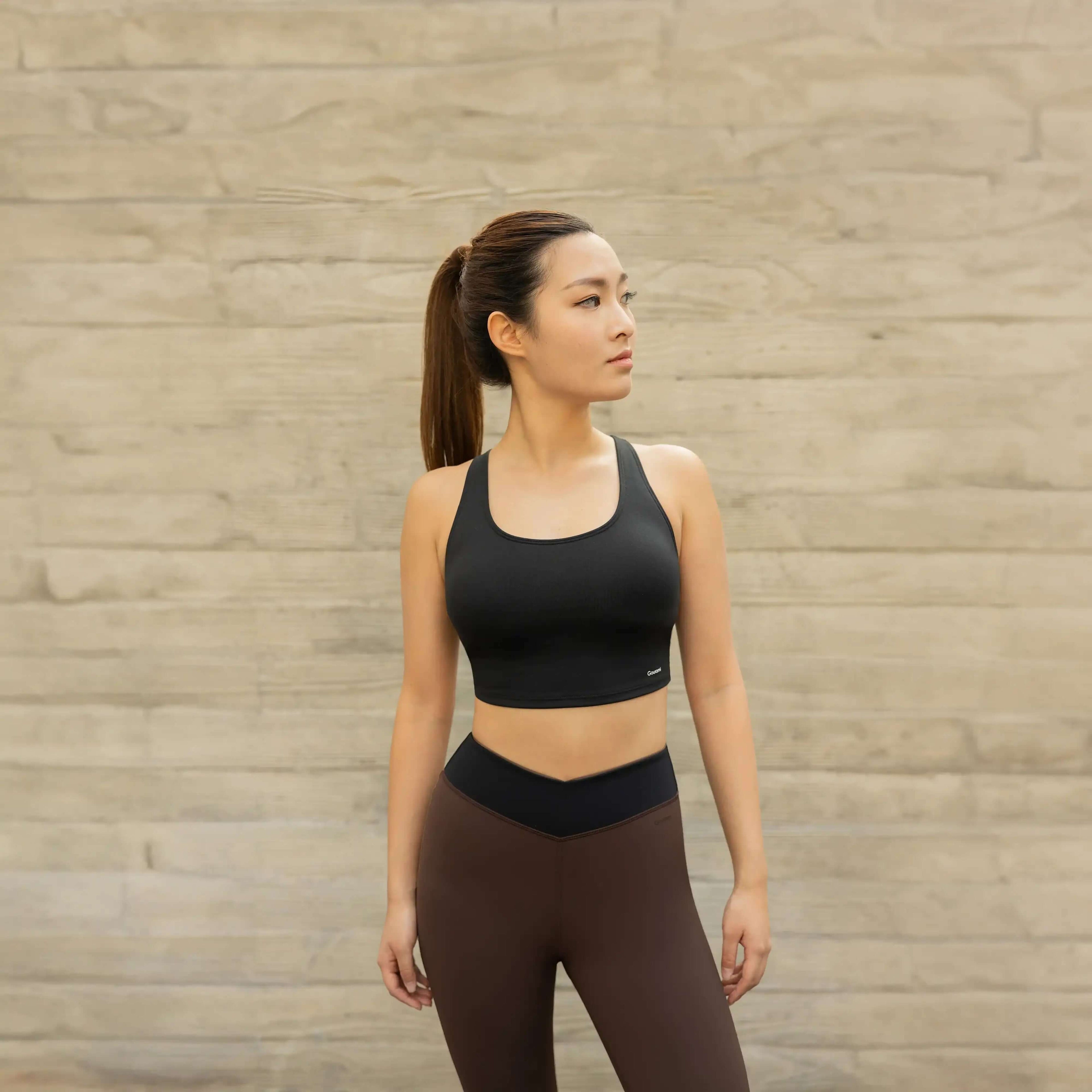 Woman in black sports bra and brown leggings, activewear against neutral wall background
