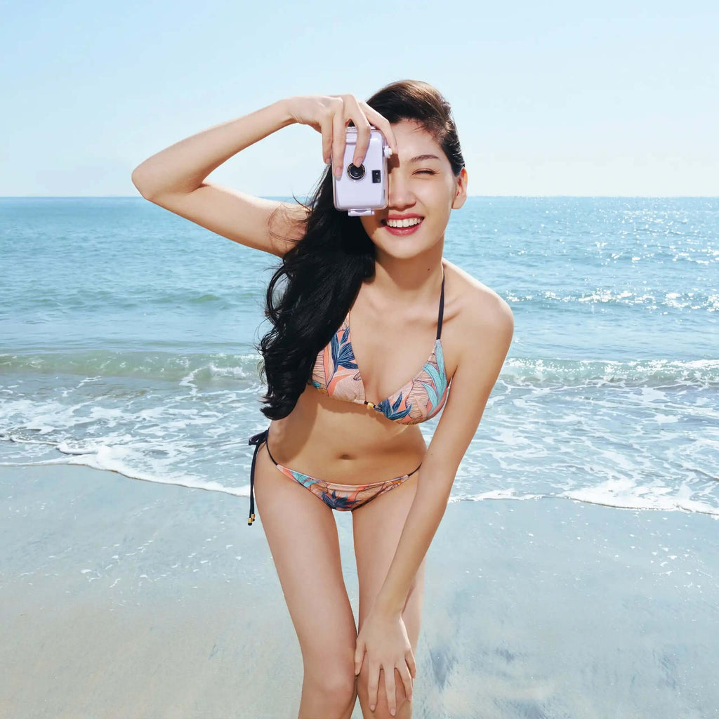 Woman in tropical print bikini at beach, holding camera, smiling under sunny sky