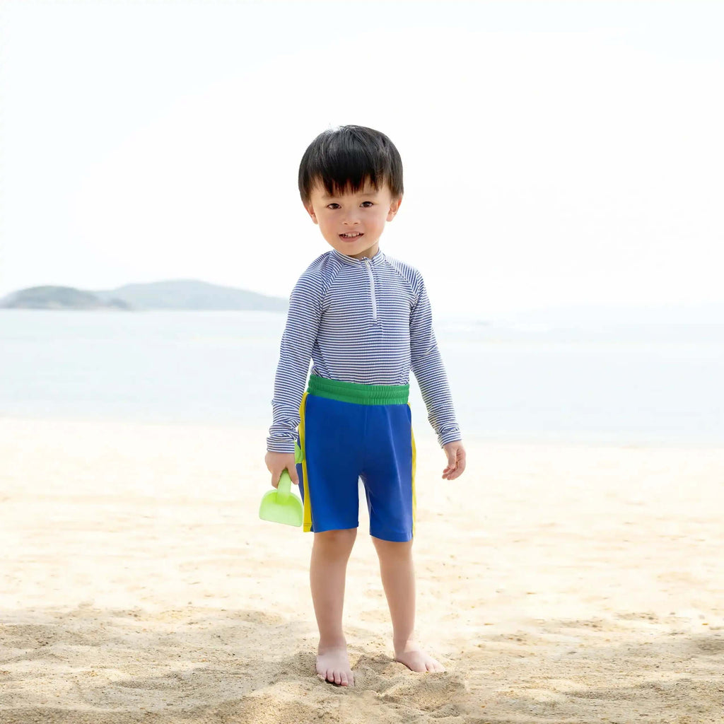 男孩在海滩上穿着糖果条纹男童Rashguard Set，在岸边拿着一个绿色水桶。