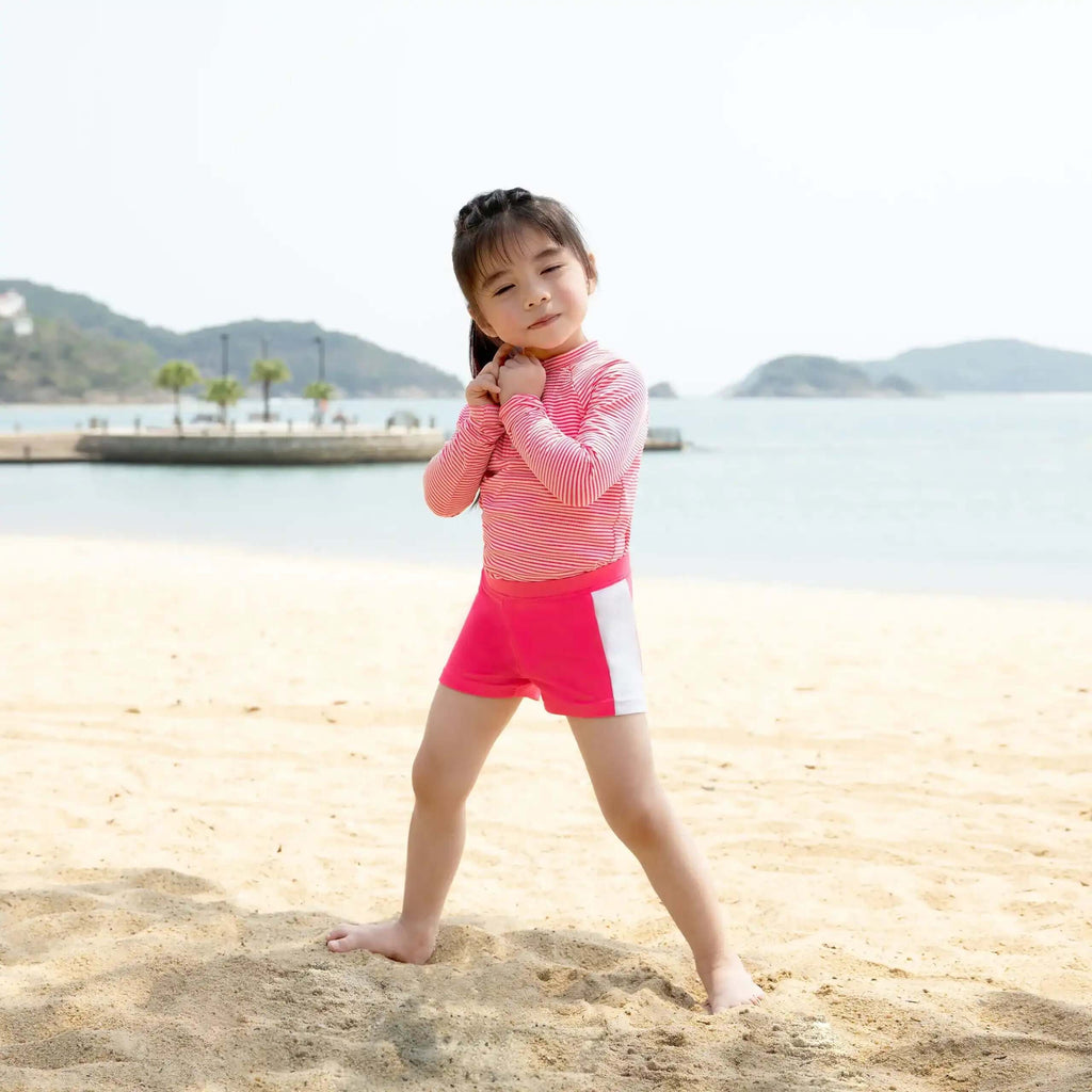 Cute girl wearing a Candy Stripe Girls Rashguard Set at the beach with a sunny backdrop.