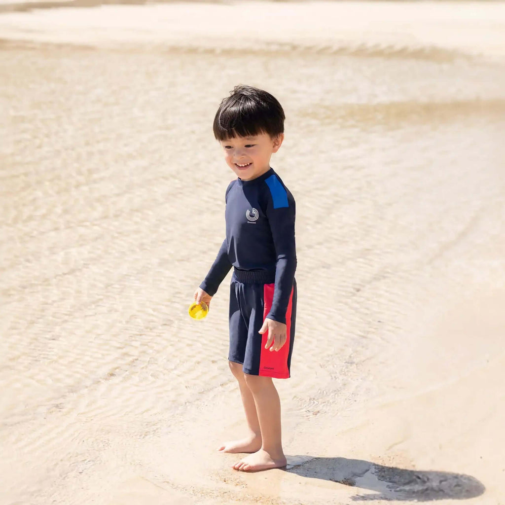 Young boy in navy swimwear with red accents standing on a sandy beach near shallow water