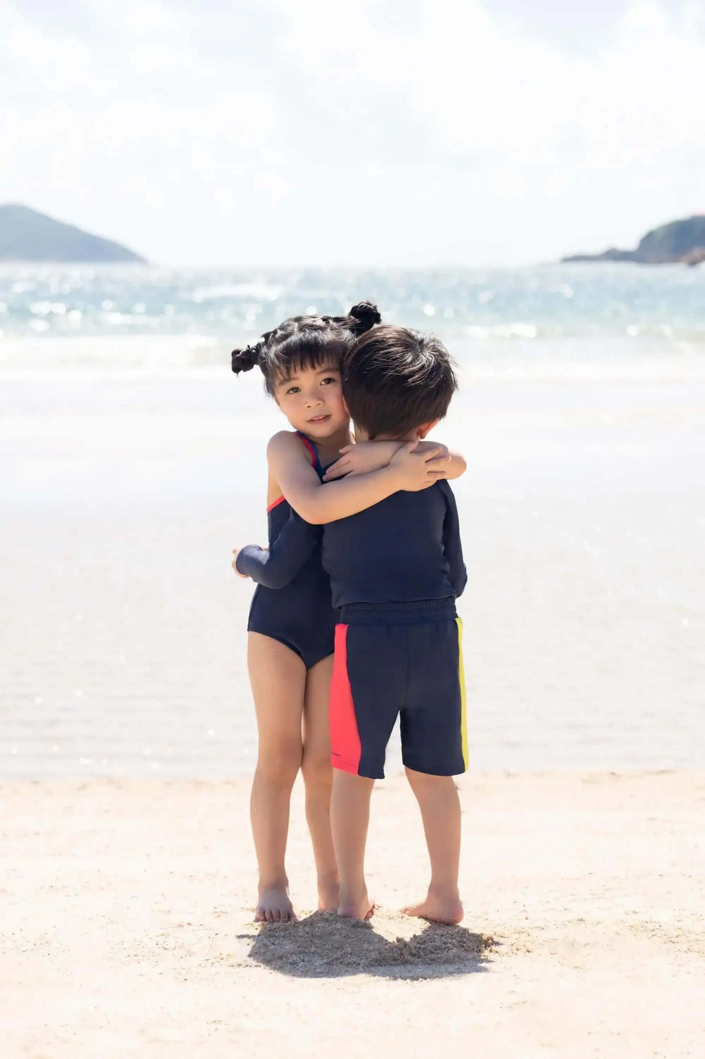 "两个穿着泳衣的孩子在阳光明媚的海滩上拥抱，背景是海浪。"