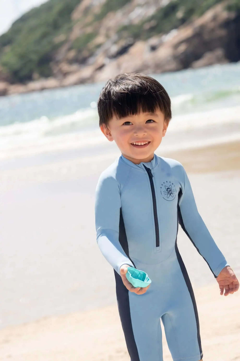 穿着浅蓝色长袖泳衣的小孩在海滩上微笑，手里拿着一个小物件
