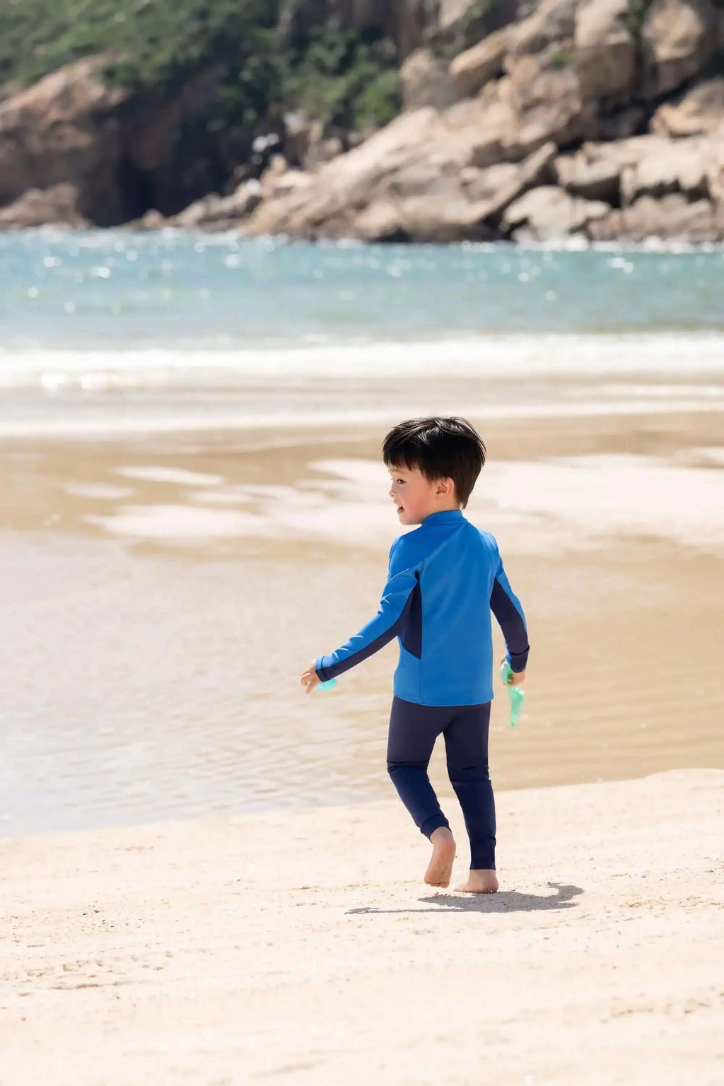 Young boy in blue long sleeve swimwear walking on sandy beach by the ocean