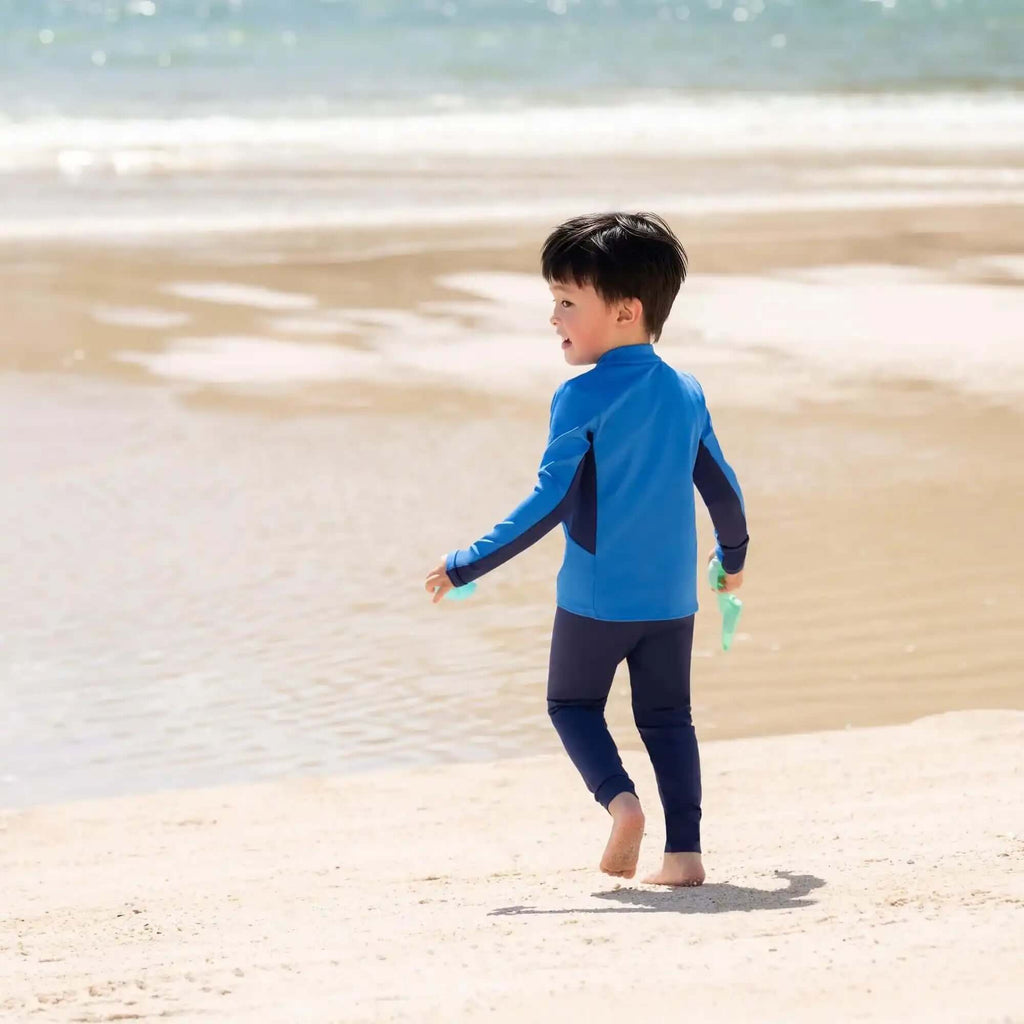 Young boy in blue swimwear walking barefoot on sandy beach near the ocean