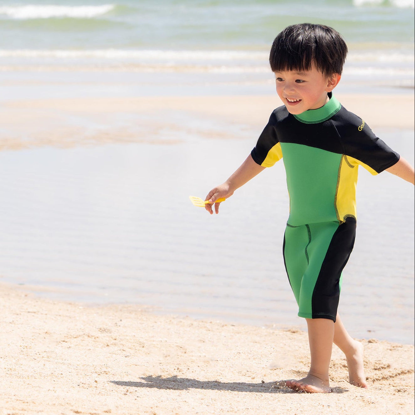 Two children in wetsuits playing on a sandy beach with ocean and sky in the background.