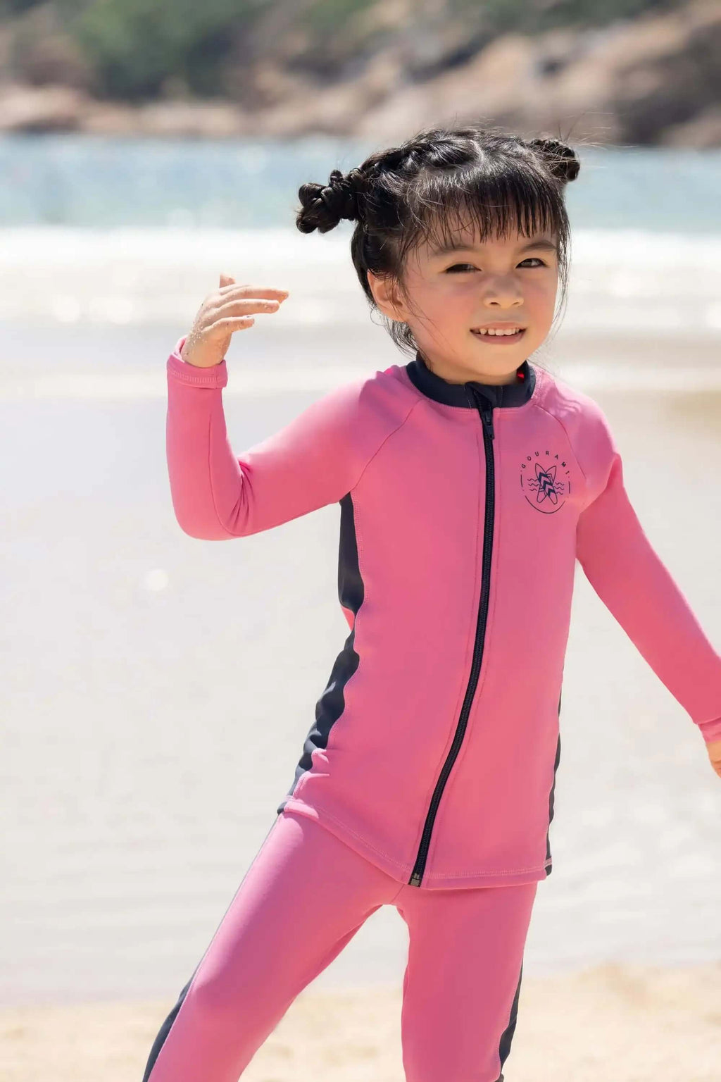 Smiling child wearing a pink zip-up rash guard set on the beach with ocean in background